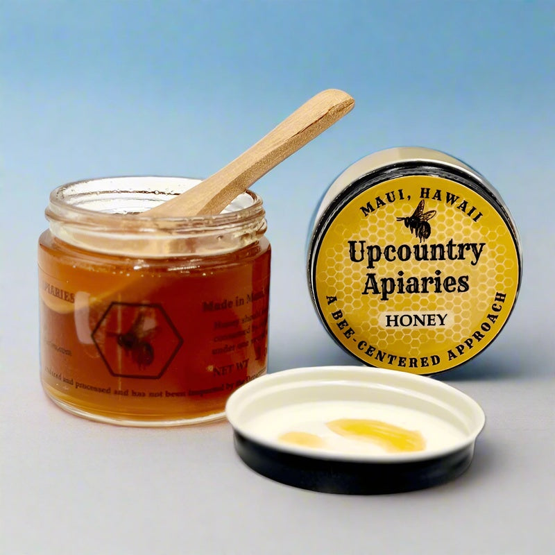 Jar of honey with a wooden spoon and Upcountry Apiaries container on a light blue background
