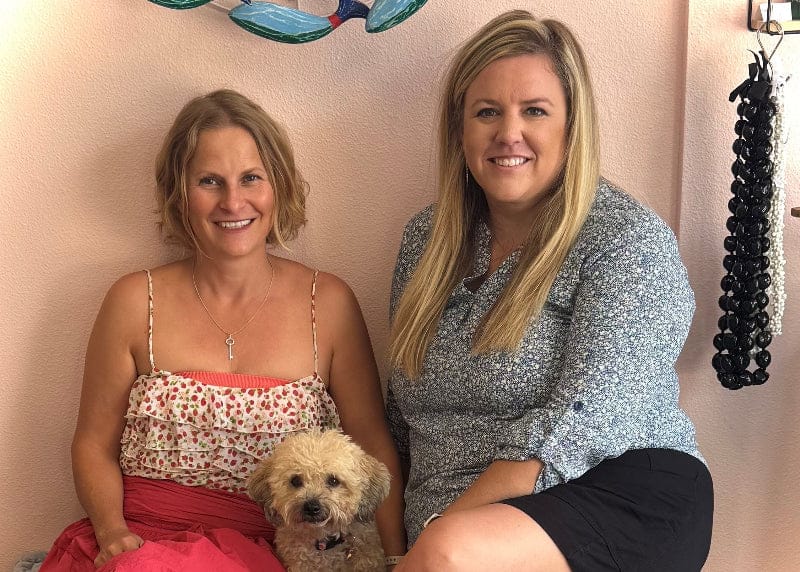 Two women sitting with a small dog against a pink wall with decorative elements.