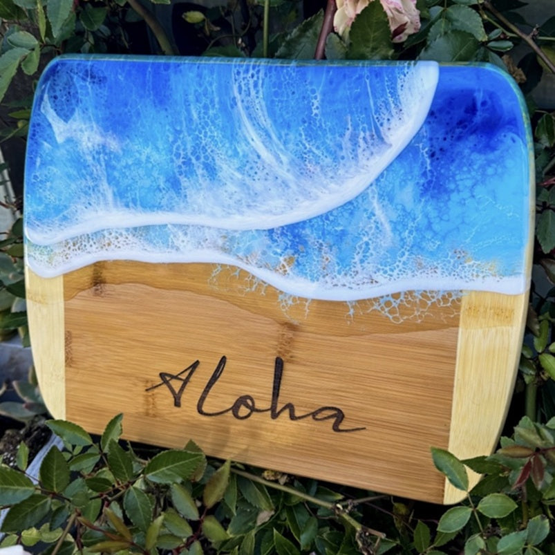 Wooden cutting board with ocean design and 'Aloha' engraving, surrounded by greenery.