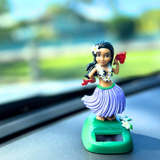 Hula dancer figurine on a car dashboard with a blurred outdoor background