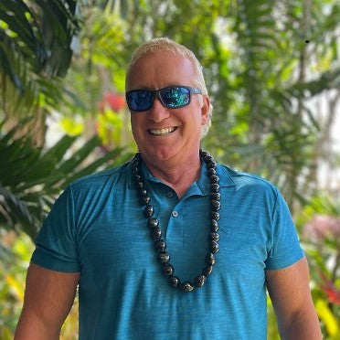 A person wearing a blue shirt and sunglasses smiling, standing in front of tropical foliage, with a kukui nut lei around their neck.