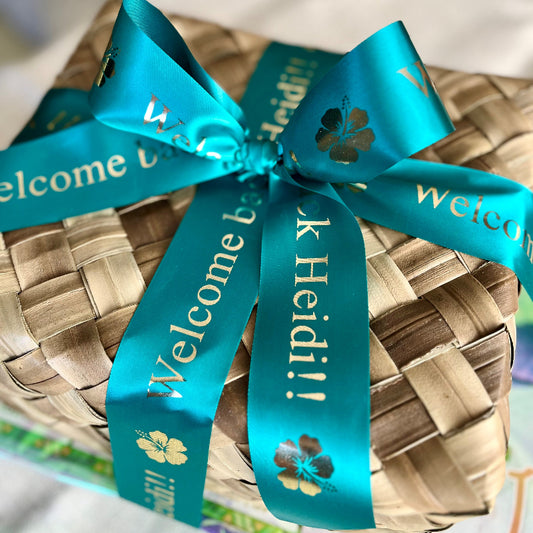 A woven lauhala gift basket with a teal ribbon that has 'Welcome to Maui' printed on it.
