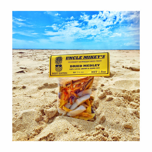 A package of Uncle Mikey's Dried Medley containing dried pineapple and other fruits, placed on a sandy beach with a bright blue sky in the background.